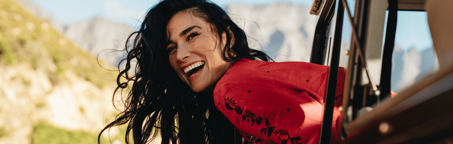 Women wearing a red blouse with black details hang outside her car window, smiling and looking happy. It's sunny outside with a mountain landscape behind her 