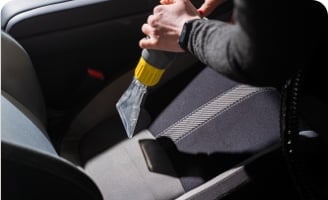 A person using a handheld vacuum on the front passenger seat of a car focused on maintaining the appearance of the car’s interior.