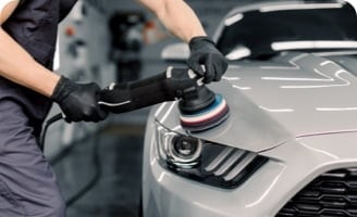 A person using a polisher on the hood of a silver car in a workshop focused on maintaining the appearance of the car’s exterior.